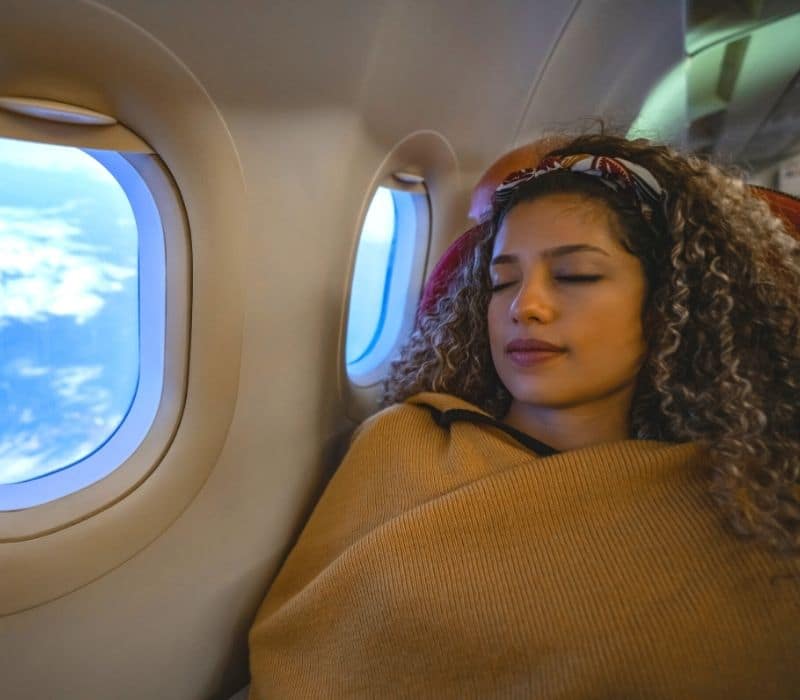 mujer que viaja sola envuelta en una manta color canela y durmiendo en un vuelo