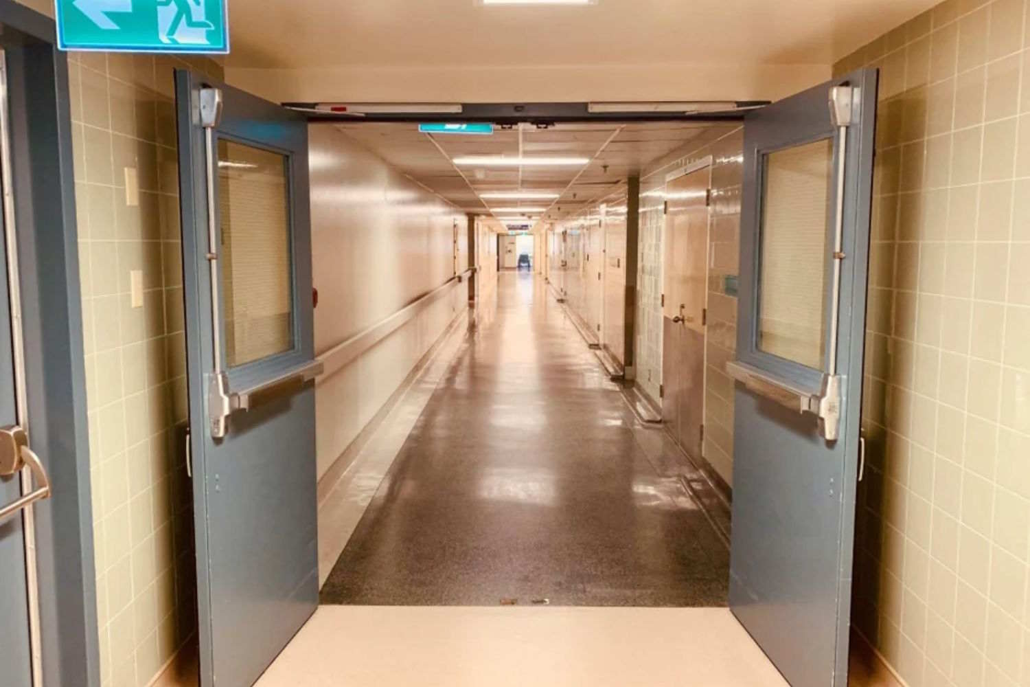 Perspectiva de un largo pasillo iluminado del hospital, enmarcado a través de puertas cortafuegos de vidrio con barras antipánico